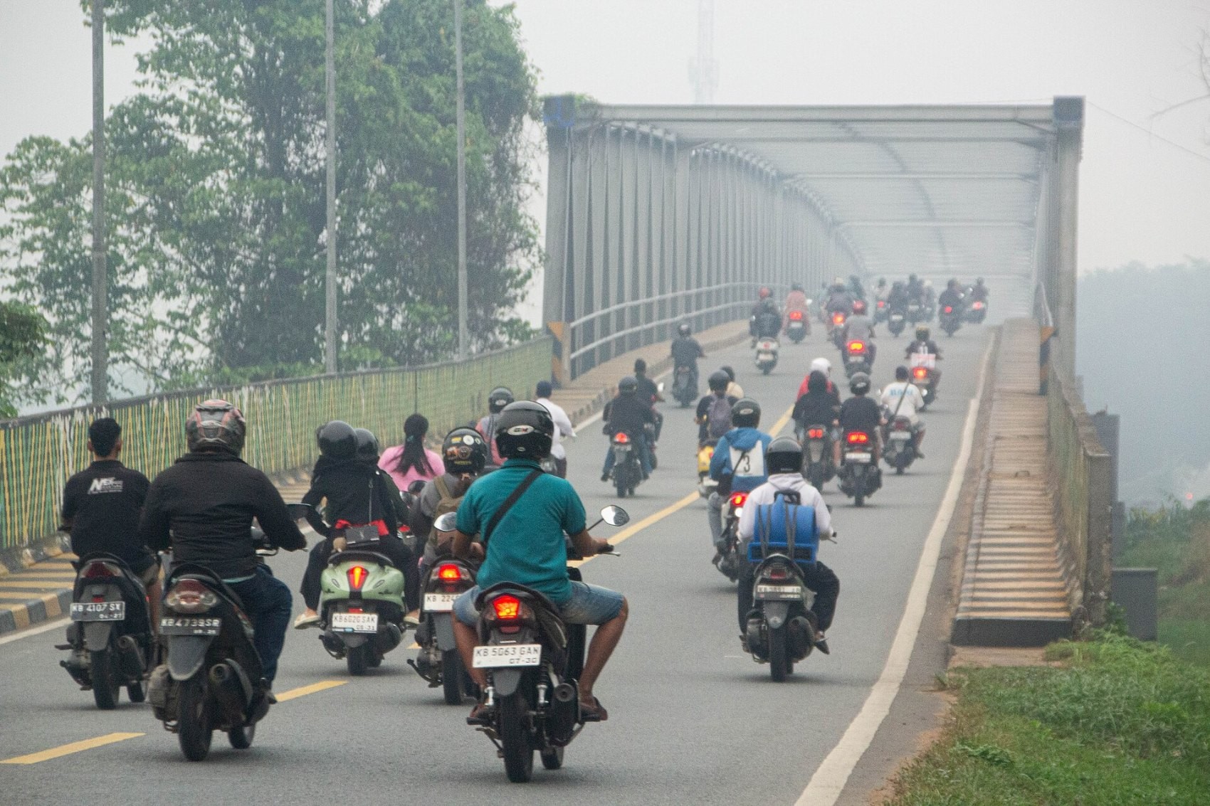 Motorcyclists riding across a bridge in Pontianak through thick grey haze.