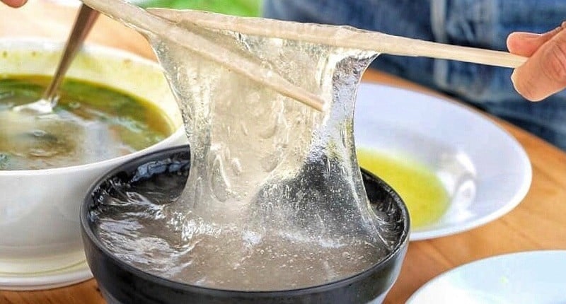 Translucent, sticky papeda being stretched upwards with chopsticks out of a dark bowl.